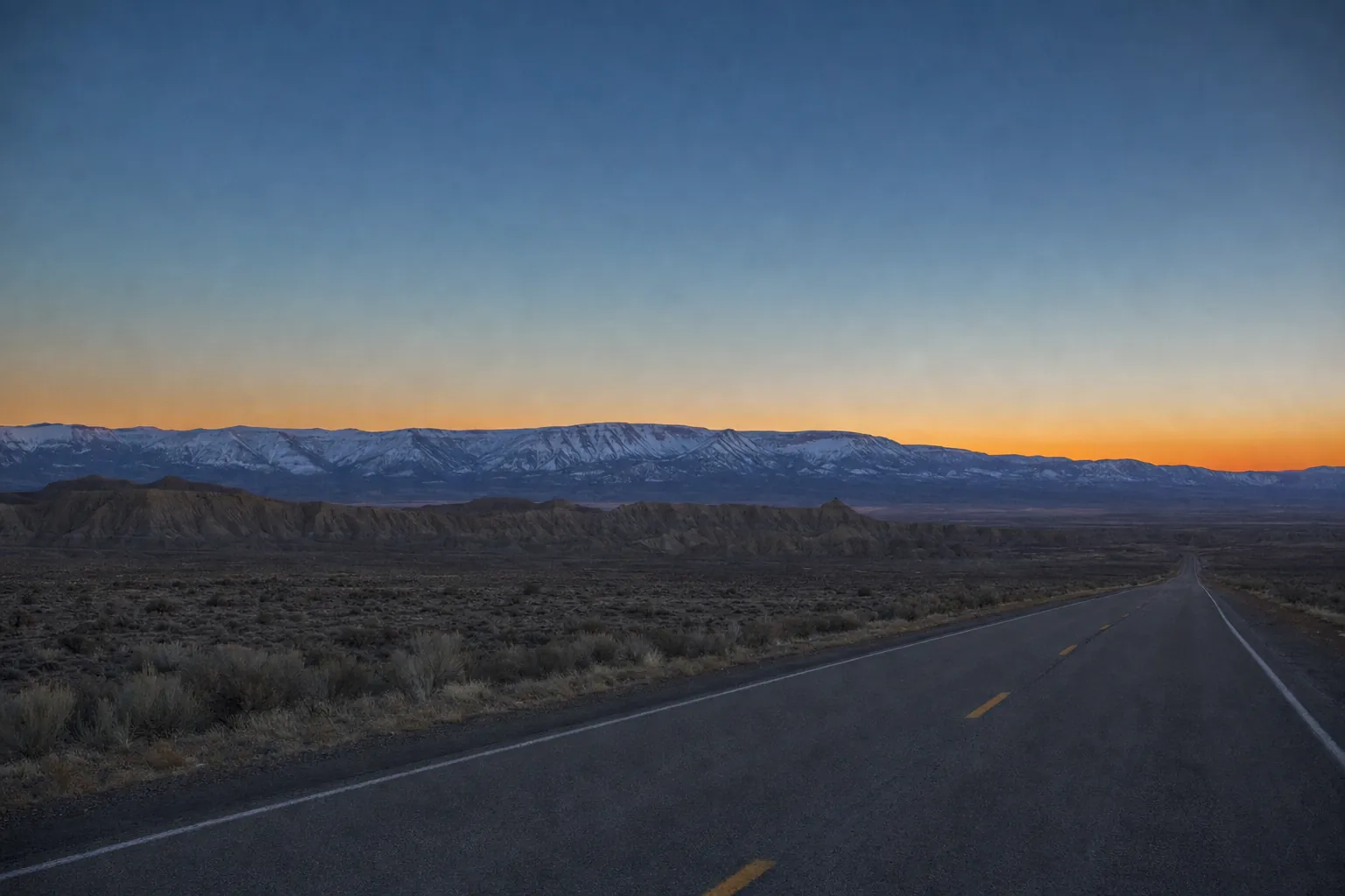 An empty two-lane highway at dusk crossing sagebrush flats, with snow-covered mountains under an orange horizon.