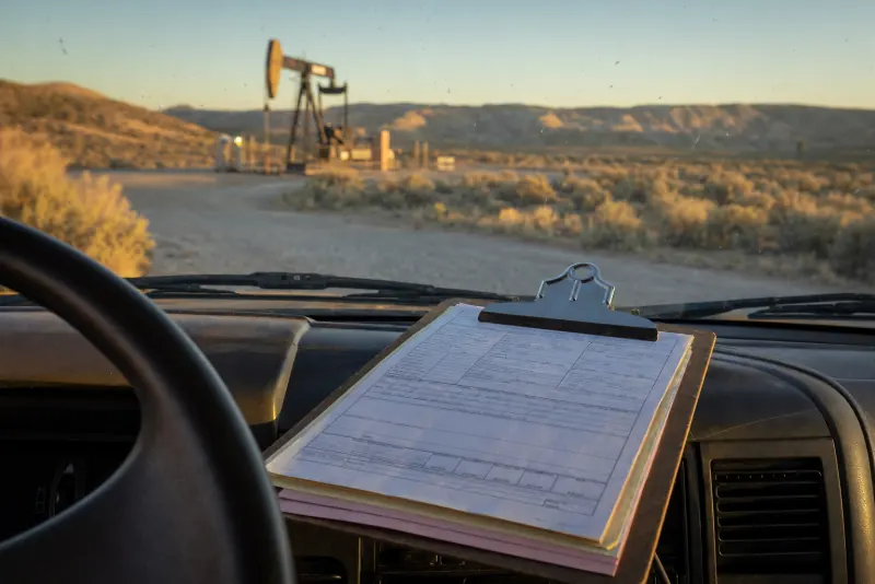A clipboard of paper tickets on a pickup dashboard, with a pumpjack and sagebrush flats through the windshield at golden hour.